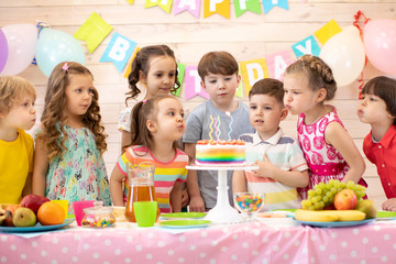 Happy children at table celebrating birthday holiday. Kids blows together candles on festive cake