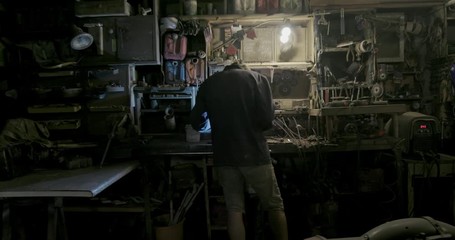 Back view of anonymous mechanic welding metal part then lifting protective mask and cooling detail in cup on workbench in garage