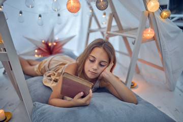 Little 10 year old girl reading classic book under her home-made tent inside the living room.