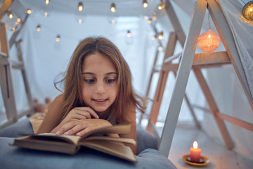 Little 10 year old girl reading classic book under her home-made tent inside the living room.
