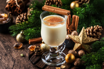 Obraz na płótnie Canvas New Year or Christmas Eggnog cocktail - hot winter or autumn drink with milk, eggs and dark rum, sprinkled with cinnamon and nutmeg in a glass on wooden background, festive decoration