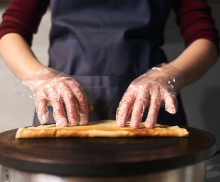Cropped view of professional cook in apron while baking crepe process on hot round cooktop. Skillful hands in gloves rolling up fried crepe. Traditional restaurant healthy food. Close up. Front view.