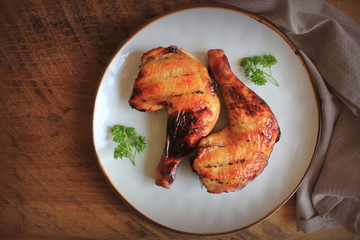 Grilled chicken leg quarters with crispy golden brown skin, lemon, parsley on white plate on dark wooden boards. Food background. Top view