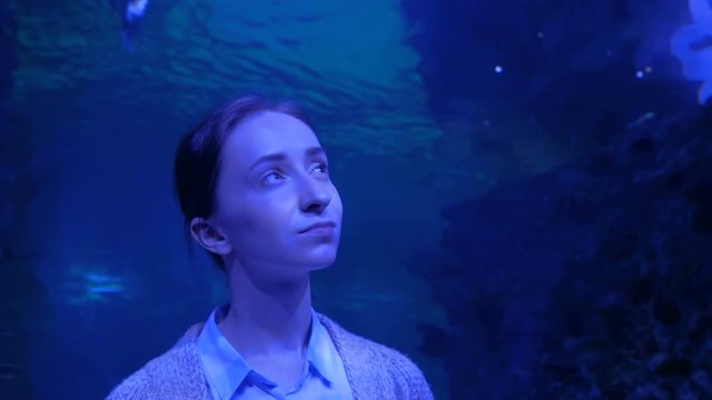 Portrait of woman looking at fish in large public aquarium tank at Oceanarium with blue illumination. Tourism, education, underwater life and entertainment concept