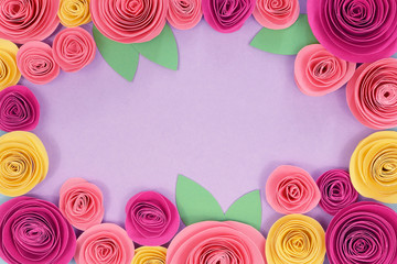 Colorful pink and yellow rose flat lay background with crafted paper flowers around the endges and empty violet copy space in middle