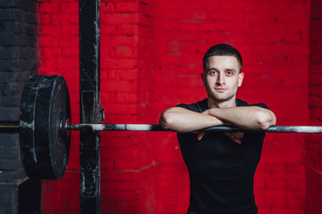 Fototapeta na wymiar Young good guy is engaged in sports. During this time he is in the gym. Dressed in a black T-shirt. Put your hands on the barbell. Look into the camera.
