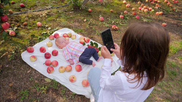 Elder sister kid girl takes mobile phone pictures of infant baby lying in apples on rug
