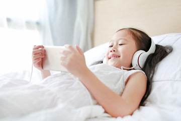 Happy little Asian girl using headphones listen music by smartphone smiling while lying on bed at home.