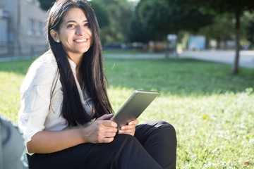 Obraz premium portrait of a beautiful indian girl. Student sitting on green grass, with backpack, smiling, holding tablet in hands
