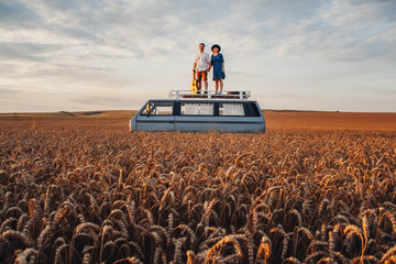 Young couple man with a guitar and woman in a hat are standing on the roof of a car in a wheat field. Travel and adventur - Powered by Adobe