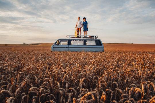 Young couple man with a guitar and woman in a hat are standing on the roof of a car in a wheat field. Travel and adventur