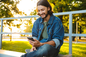 Happy smiling young bearded man with skateboard sit in nature park using mobile phone. © Drobot Dean