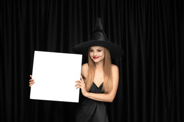 Young blonde woman in black hat and costume on black background. Attractive, sensual female model. Halloween, black friday, cyber monday, sales, autumn. Holding white sheet for copyspace, looks mystic