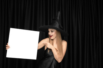 Young blonde woman in black hat and costume on black background. Attractive, sensual female model. Halloween, black friday, cyber monday, sales, autumn. Holding white sheet for copyspace, looks mystic