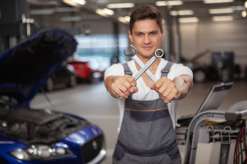 Cheerful auto mechanic holding out two wrenches to the camera. Young male auto service worker holding crossed tools. Car service concept