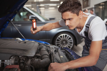 Fototapeta na wymiar Young male car service worker looking under the hood of an automobile at the garage. Handsome auto mechanic examining engine of an auto at his workshop
