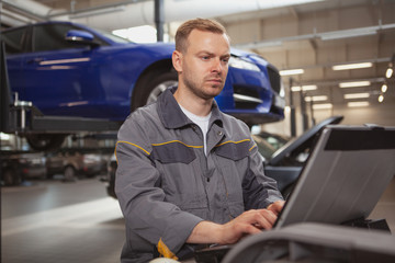 Mature male car mechanic looking concentrated, doing computer diagnostics on a broken automobile at his workshop. Cars, transportation concept