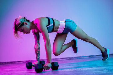 Athletic young blonde woman doing heavy dumbbell exercise in the gym in neon lights.