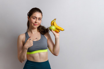 Sport and health. Beautiful athletic young woman in gym clothes with Apple and banana in her hands pointing at them with her finger. Copy space