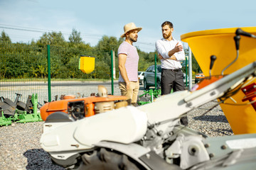 Young agronomist with salesman at the open ground of the shop with agricultural machinery, buying a new planter for farming