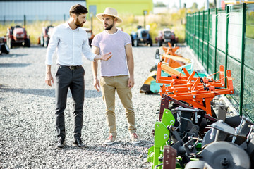 Young agronomist walking with salesman at the open ground of the shop with agricultural machinery,...