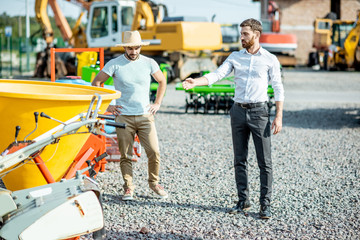 Fototapeta premium Young agronomist with salesman at the open ground of the shop with agricultural machinery, buying a new planter for farming