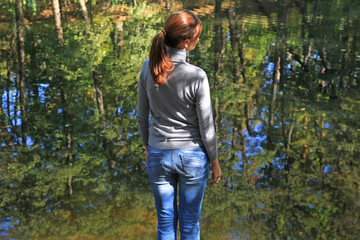 Young girl is standing near the pond and looking into the distance. Autumn morning landscape with woman and sun rays that reflected on the clear water. Pastime outdoors