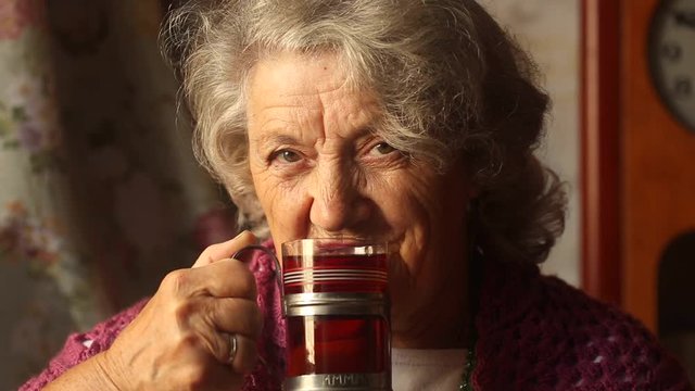 Elderly happy woman smile and drink tea from glass and old cup holder on a dark background