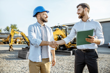 Builder choosing heavy machinery for construction with a sales consultant shaking hands on the open...