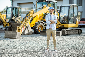 Portrait of a handsome builder standing on the open ground of the shop with heavy machinery for...