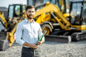Fotobehang Vrachtwagen Portrait of a handsome sales consultant or manager standing on the open ground of the shop with heavy machinery for construction  © rh2010