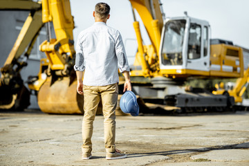 Portrait of a handsome builder standing back on the open ground of the shop with heavy machinery for construction