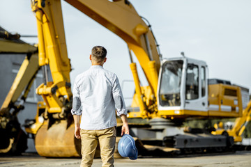 Portrait of a handsome builder standing back on the open ground of the shop with heavy machinery for construction