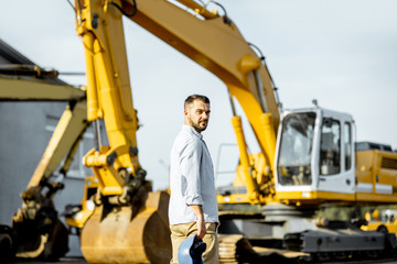 Portrait of a handsome builder standing on the open ground of the shop with heavy machinery for construction