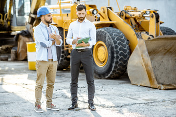 Builder choosing heavy machinery for construction with a sales consultant, signing some documents on the open ground of a shop with special vehicles