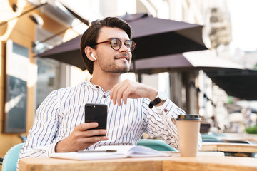 Photo of smiling caucasian man using earpod and cellphone while working in cafe outdoors