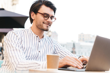 Photo of cheerful caucasian man typing on laptop while drinking coffee in cafe outdoors