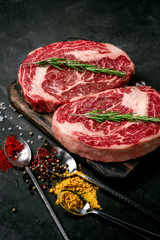 Two raw rib eye steak with seasonings on the dark stone background prepared for cooking. Marbled beef top view