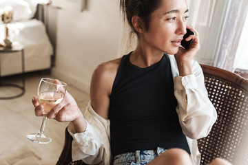 Portrait of charming woman talking on smartphone and drinking wine while sitting on armchair in apartment