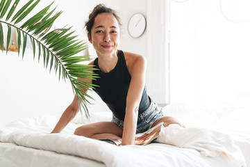 Portrait of caucasian woman smiling while reading magazine on bed in bright room with green plant