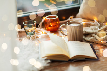 hygge and cozy home concept - book, cup of coffee or hot cchocolate and candles with garland on...