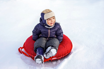 happy joyful beautiful child boy rides from the mountain on a red tubing in winter