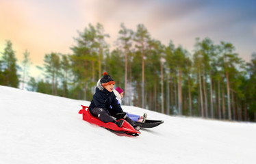 Fototapeta na wymiar childhood, sledging and season concept - happy little kids sliding on sleds down snow hill over winter forest background