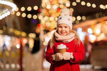 holidays, childhood and people concept - happy little girl with cup of tea at christmas market in winter evening