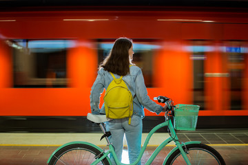 Teenager girl in jeans with yellow backpack and bike standing on metro station, waiting for train, smiling, laughing. Orange train passing by behind the girl. Futuristic subway station. Finland, Espoo © Suzi Media 