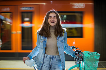 Teenager girl in jeans with yellow backpack and bike standing on metro station, waiting for train,...