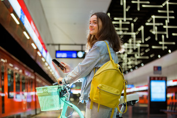 Teenager girl with backpack and bike standing on metro station holding smart phone in hand, scrolling and texting, smiling and laughing. Futuristic bright subway station. Finland, Espoo