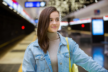 Teenager girl in jeans with yellow backpack and bike standing on metro station, waiting for train, smiling and laughing. Futuristic bright subway station. Finland, Espoo