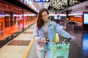 Teenager girl in jeans with yellow backpack and bike standing on metro station, waiting for train, smiling and laughing. Futuristic bright subway station. Finland, Espoo