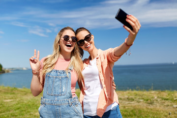 leisure and friendship concept - happy smiling teenage girls or best friends in sunglasses hugging and taking selfie by smartphone at seaside in summer